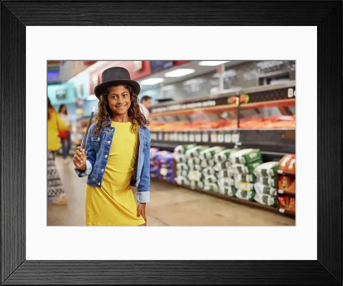 Framed Print : Young girl with magicians top hat and wand standing in isle of grocery store, meat market in the background