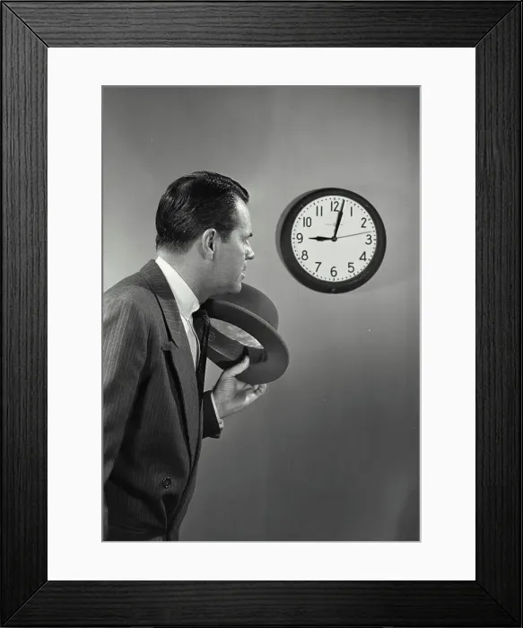 Framed Print : Vintage photograph. Man with dark hair and suit holding hat standing next to wall clock