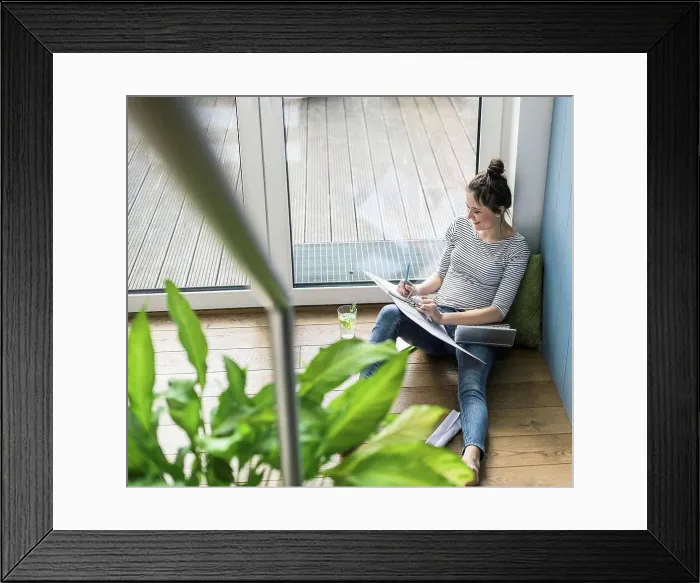 Framed Print : Bird's eye view of smiling woman sitting at the window at home working with laptop and file folder