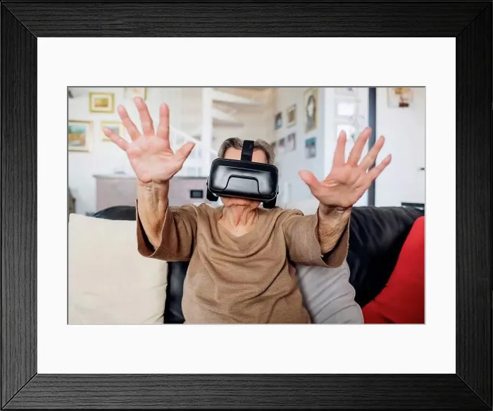 Framed Print : Senior woman wearing virtual reality simulator gesturing while siting on sofa in living room at home