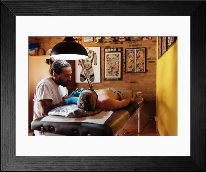 Framed Print : Side view of tattoo artist working on a customer's arm in his studio Framed Print : Side view of tattoo artist working on a customer's arm in his studio