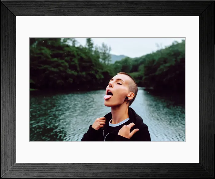 Framed Print : Short haired female with piercing wearing black hoodie looking up while catching raindrops with tongue near green Framed Print : Short haired female with piercing wearing black hoodie looking up while catching raindrops with tongue near green