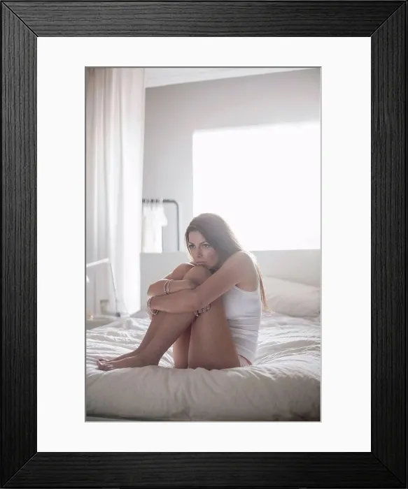 Framed Print : Young woman embracing knee to her chest while sitting on bedroom at home Framed Print : Young woman embracing knee to her chest while sitting on bedroom at home