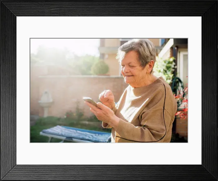 Framed Print : Smiling senior woman using smart phone while standing at backyard during sunny day Framed Print : Smiling senior woman using smart phone while standing at backyard during sunny day
