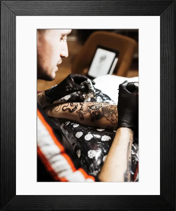Framed Print : From above stylish man with piercing using tattoo machine to make tattoo on leg of crop customer during work in salon