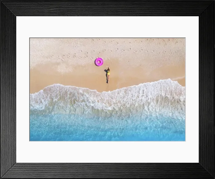 Framed Print : Aerial view of the lying beautiful young woman with pink swim ring on the sandy beach near sea with waves at sunset Framed Print : Aerial view of the lying beautiful young woman with pink swim ring on the sandy beach near sea with waves at sunset