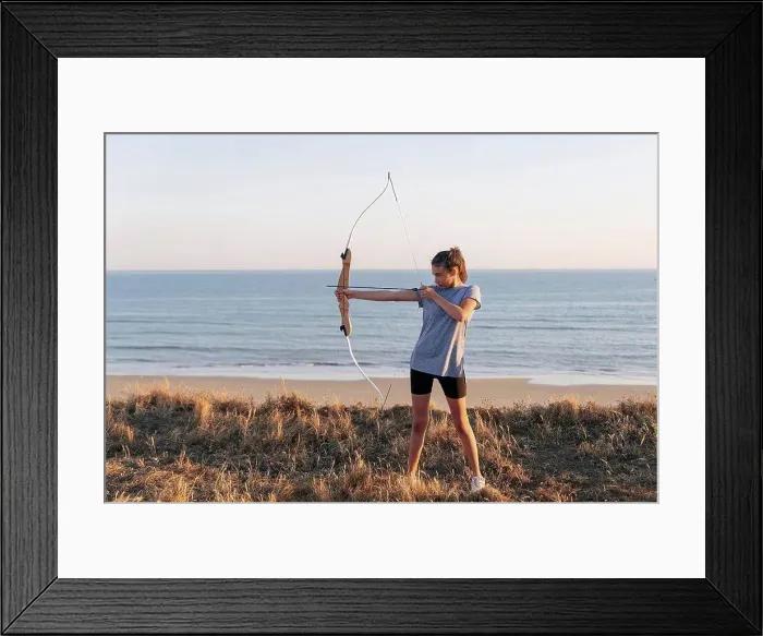 Framed Print : Archeress aiming with bow and arrow while standing on grass at beach