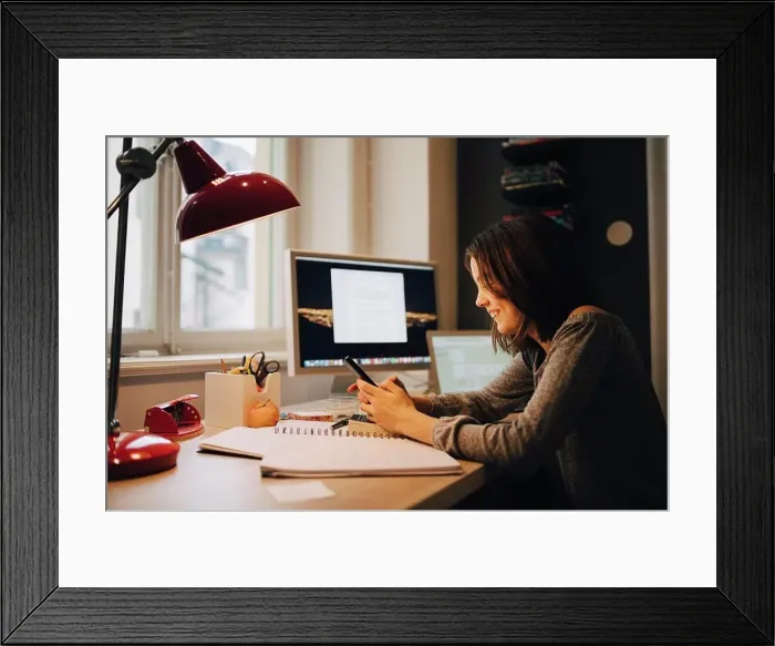 Framed Print : Happy girl using mobile phone while sitting at illuminated desk Framed Print : Happy girl using mobile phone while sitting at illuminated desk