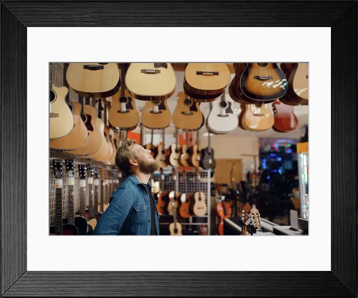Framed Print : Bearded young guy choosing acoustic guitar in music store Framed Print : Bearded young guy choosing acoustic guitar in music store