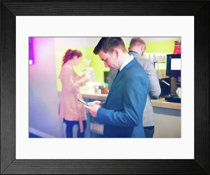Framed Print : Businessmen and women getting coffee in airport lounge, man in foreground using smartphone Framed Print : Businessmen and women getting coffee in airport lounge, man in foreground using smartphone