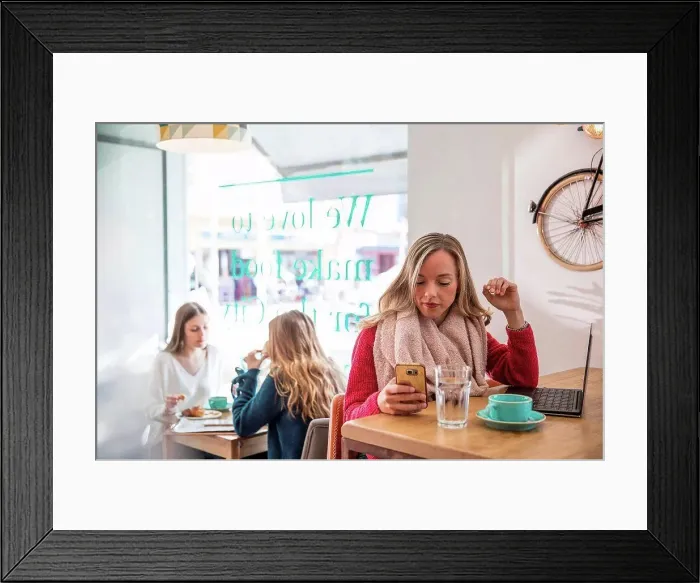 Framed Print : Woman sitting in cafe, looking at smartphone, laptop in front of her Framed Print : Woman sitting in cafe, looking at smartphone, laptop in front of her