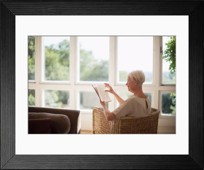 Framed Print : Senior woman relaxing and reading book in sunny living room