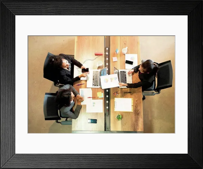 Framed Print : Three businesswomen sitting at desk, using laptop, looking at smartphone, overhead view Framed Print : Three businesswomen sitting at desk, using laptop, looking at smartphone, overhead view