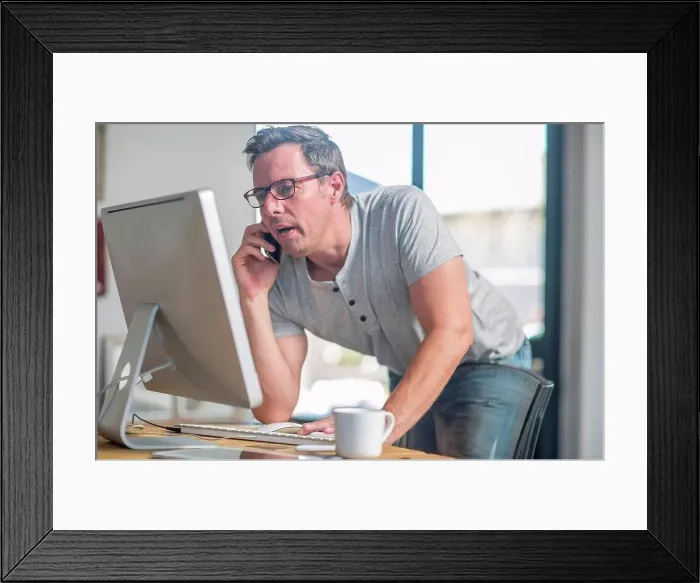 Framed Print : Man at computer using smartphone to make telephone call Framed Print : Man at computer using smartphone to make telephone call