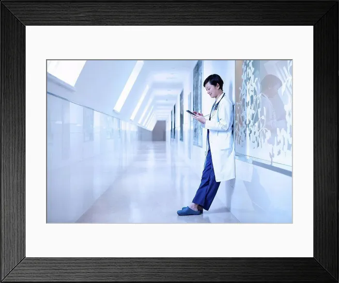 Framed Print : Doctor in hospital corridor leaning against wall using smartphone Framed Print : Doctor in hospital corridor leaning against wall using smartphone