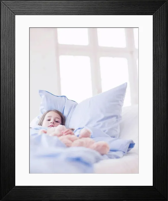 Framed Print : A young girl with brown hair lying in bed holding on to a teddy bear looking at viewer Framed Print : A young girl with brown hair lying in bed holding on to a teddy bear looking at viewer