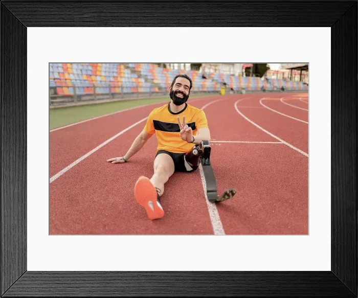 Framed Print : Smiling man with prosthetic leg sitting on running track showing peace sign Framed Print : Smiling man with prosthetic leg sitting on running track showing peace sign