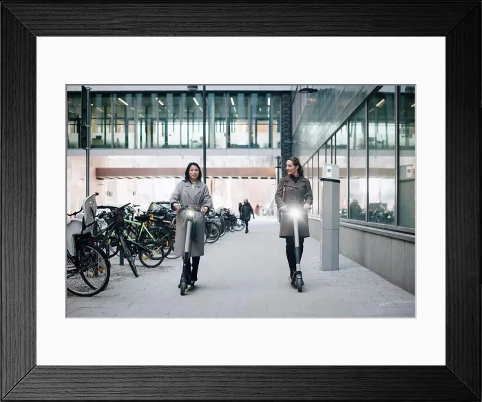 Framed Print : Full length of confident female colleagues riding electric push scooter on footpath by building in city
