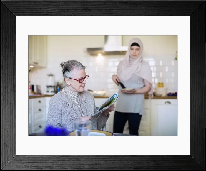 Framed Print : Senior woman reading newspaper with female home caregiver preparing food in kitchen