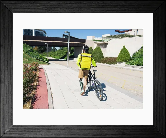Framed Print : Delivery man with prosthetic leg wheeling bicycle on footpath Framed Print : Delivery man with prosthetic leg wheeling bicycle on footpath