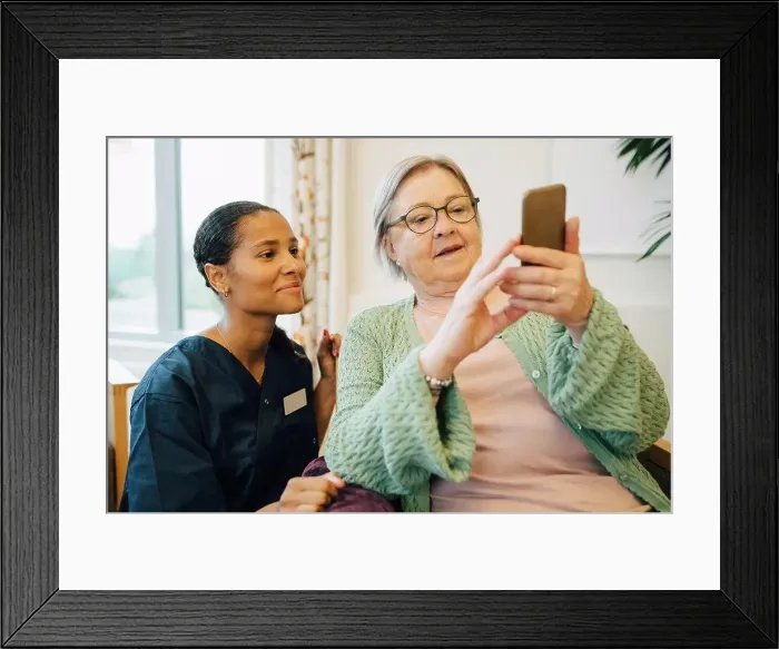 Framed Print : Smiling female caretaker looking at senior woman using mobile phone in nursing home Framed Print : Smiling female caretaker looking at senior woman using mobile phone in nursing home