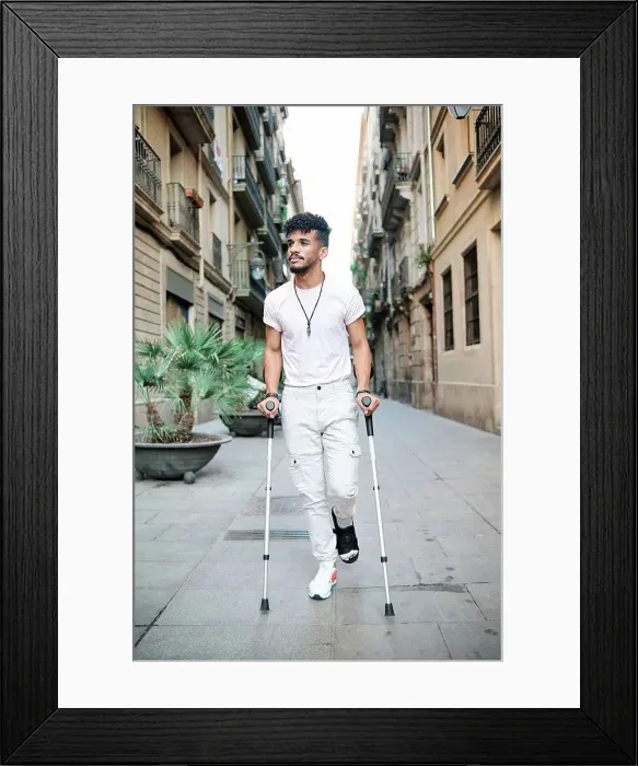 Framed Print : Young man walking in alley with help of crutches amidst buildings Framed Print : Young man walking in alley with help of crutches amidst buildings
