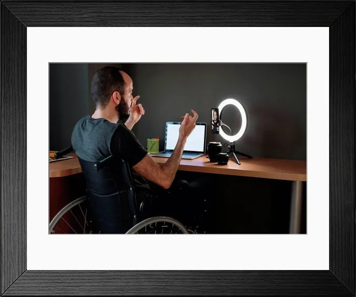 Framed Print : Vlogger filming through mobile phone with circular strobe tripod while sitting on wheelchair at studio Framed Print : Vlogger filming through mobile phone with circular strobe tripod while sitting on wheelchair at studio