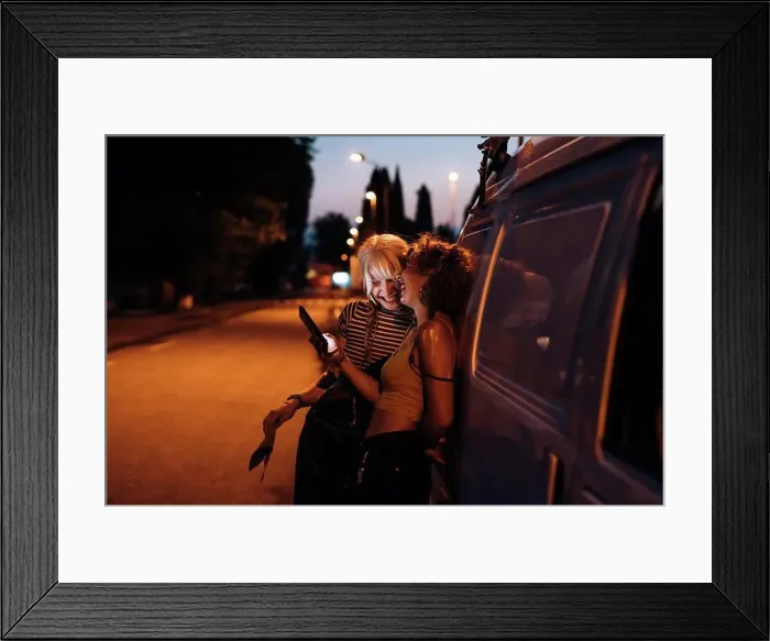 Framed Print : Female couple laughing with phone at night, leaning on van