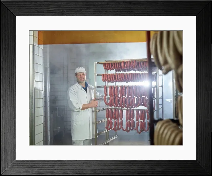 Framed Print : Smiling butcher standing by arranged sausages in smokehouse Framed Print : Smiling butcher standing by arranged sausages in smokehouse
