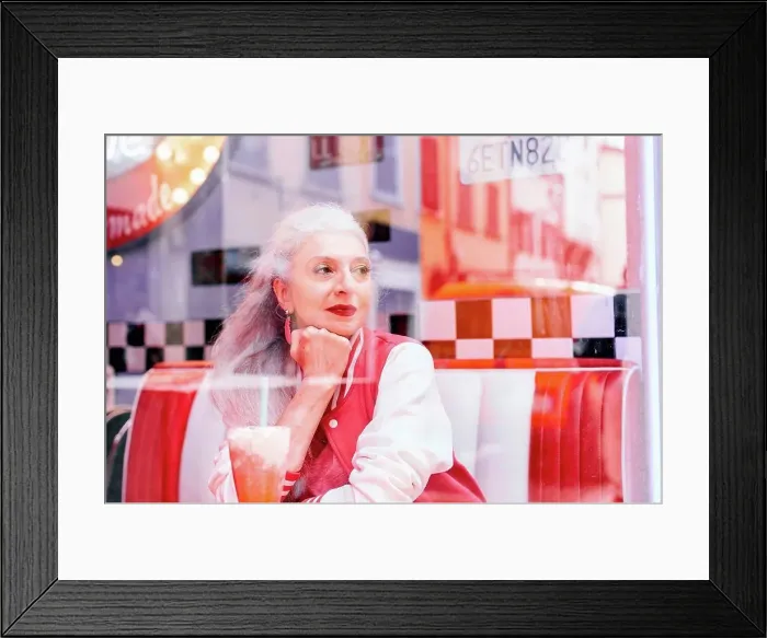 Framed Print : Mature woman in baseball jacket looking through window from 1950's diner