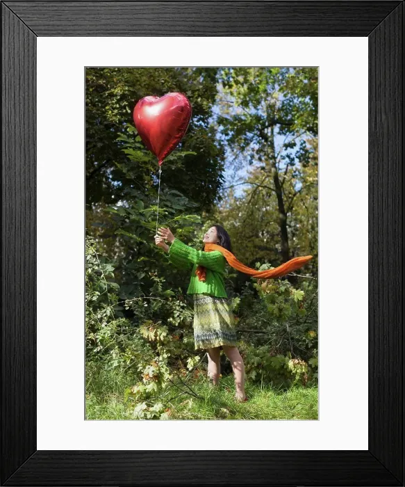 Framed Print : Smiling woman holding red heart shape balloon while standing on field against trees at park Framed Print : Smiling woman holding red heart shape balloon while standing on field against trees at park