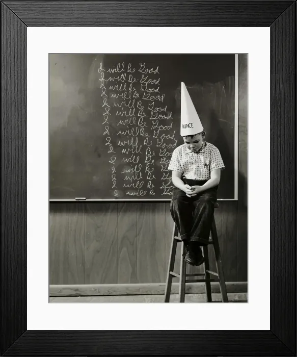 Framed Print : Boy wearing a dunce cap sitting in front of a blackboard Framed Print : Boy wearing a dunce cap sitting in front of a blackboard