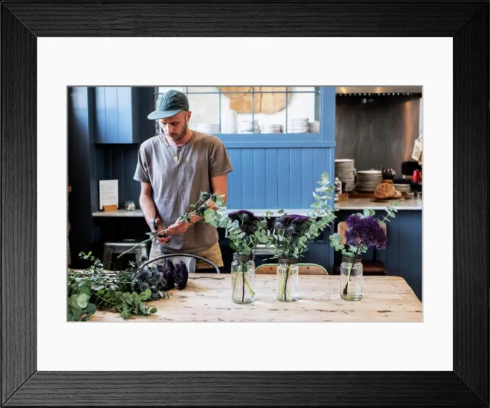 Framed Print : Man wearing baseball cap standing at a table, cutting thistles for flower arrangements in large glass jars