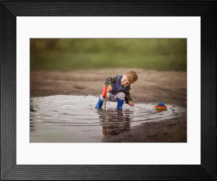 Framed Print : Boy in rain boots crouching in puddle and playing with ships Framed Print : Boy in rain boots crouching in puddle and playing with ships