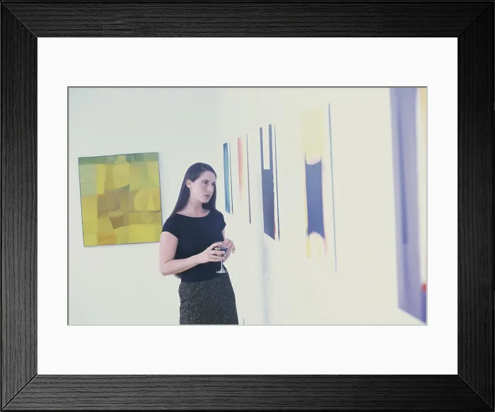 Framed Print : Young woman holding a glass looking at paintings on a wall Framed Print : Young woman holding a glass looking at paintings on a wall