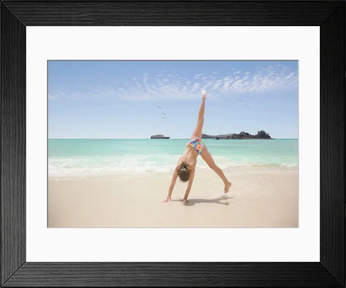 Framed Print : Teenage girl doing a cartwheel on the beach, cruise ship in ocean in background, Galapagos Islands
