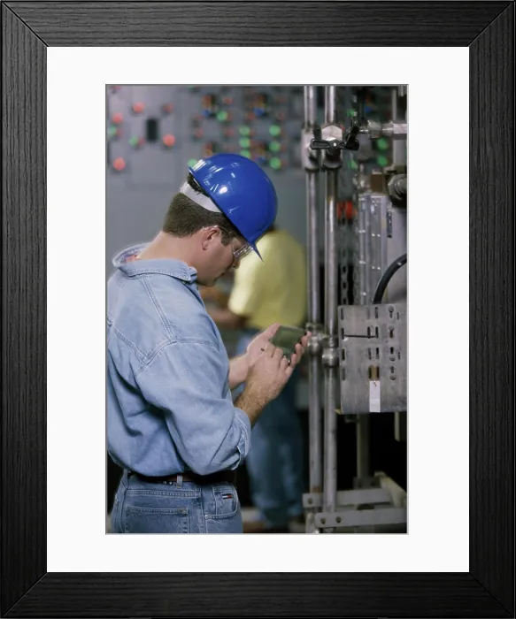 Framed Print : Worker standing beside machinery at a power plant operating a hand held device Framed Print : Worker standing beside machinery at a power plant operating a hand held device