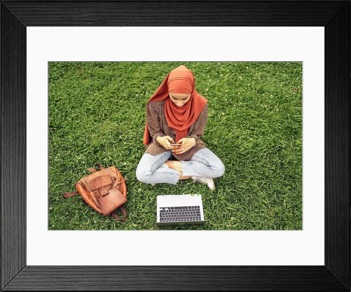 Framed Print : Muslim woman siting on the grass using a computer and phone