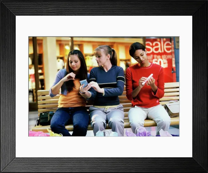 Framed Print : Three teenage girls sitting on a bench in a shopping mall applying make-up