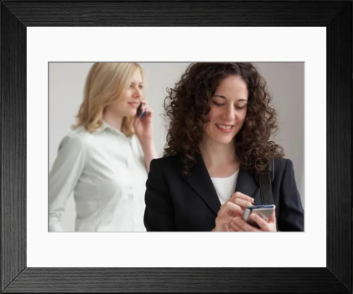 Framed Print : Close-up of a businesswoman using a PDA with another businesswoman talking on a mobile phone Framed Print : Close-up of a businesswoman using a PDA with another businesswoman talking on a mobile phone