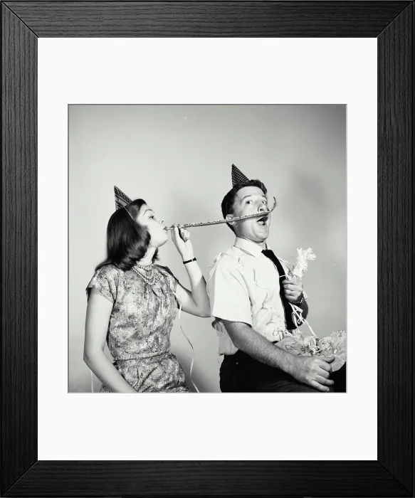 Framed Print : Young woman blowing a noisemaker near a young man's face