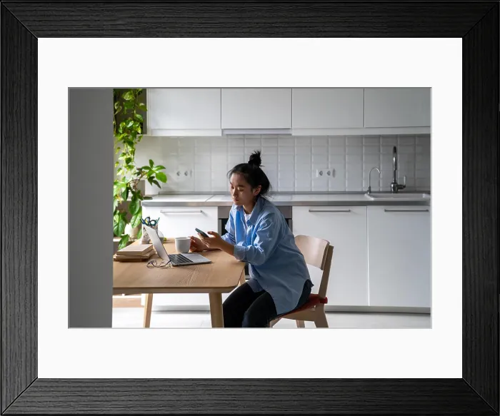 Framed Print : Asian woman sits at the kitchen table with a laptop Framed Print : Asian woman sits at the kitchen table with a laptop