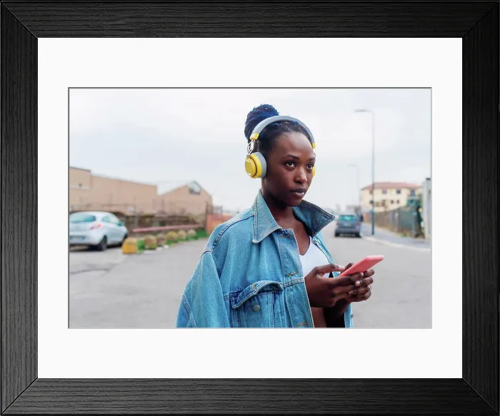 Framed Print : Italy, Milan, Woman with headphones holding smart phone in city Framed Print : Italy, Milan, Woman with headphones holding smart phone in city