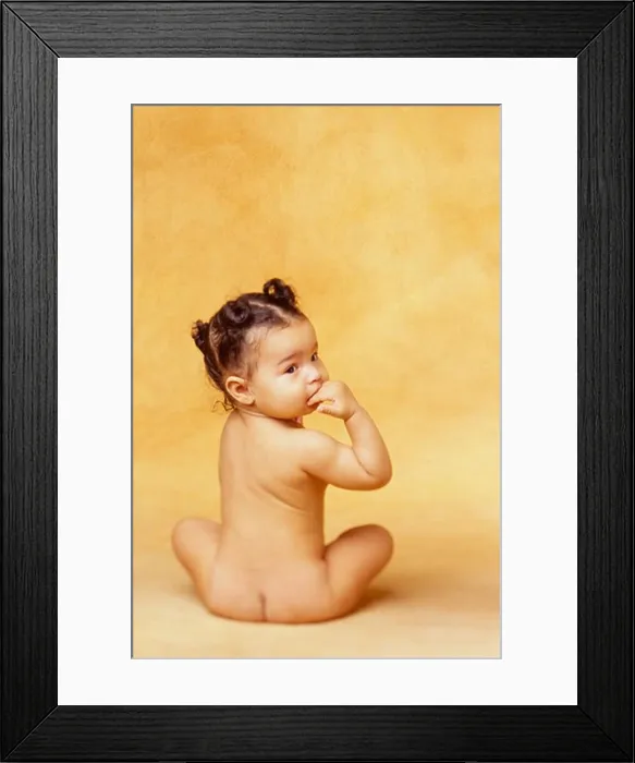 Framed Print : Naked African-American baby sits in a studio with her back to the camera and looks over her shoulder