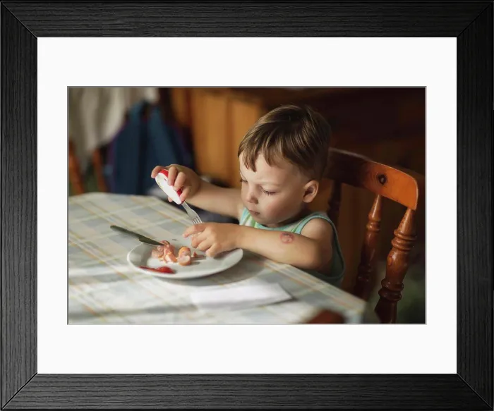 Framed Print : Boy sittyng by table and eating hot-dogs with fork Framed Print : Boy sittyng by table and eating hot-dogs with fork