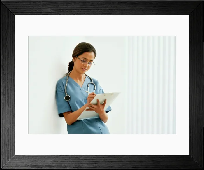 Framed Print : Young female surgeon writing notes while leaning on wall Framed Print : Young female surgeon writing notes while leaning on wall