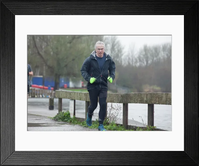 Framed Print : A mature man jogging along a waterside path on an overcast day, wearing a black jacket and bright green gloves