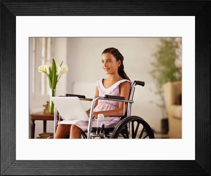 Framed Print : Young girl sitting in a wheelchair looks up from a laptop on her lap and smiles Framed Print : Young girl sitting in a wheelchair looks up from a laptop on her lap and smiles