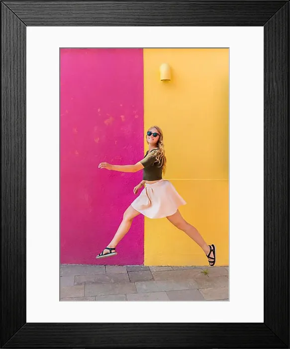 Framed Print : Young woman in dress jumps happily in front of colorful house Framed Print : Young woman in dress jumps happily in front of colorful house