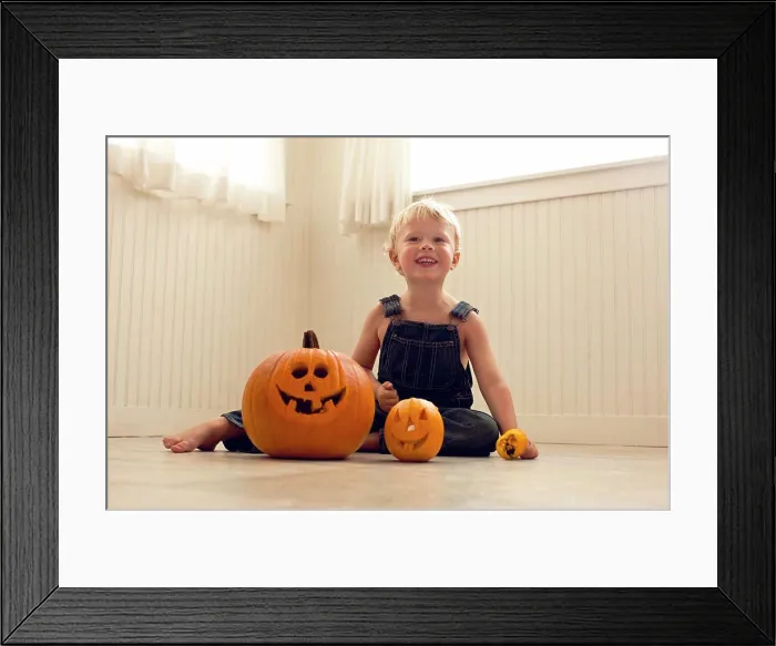 Framed Print : Smiling, young boy sits a floor behind a big Jack O'Lantern and a small Jack O'Lantern as he poses for a portrait Framed Print : Smiling, young boy sits a floor behind a big Jack O'Lantern and a small Jack O'Lantern as he poses for a portrait